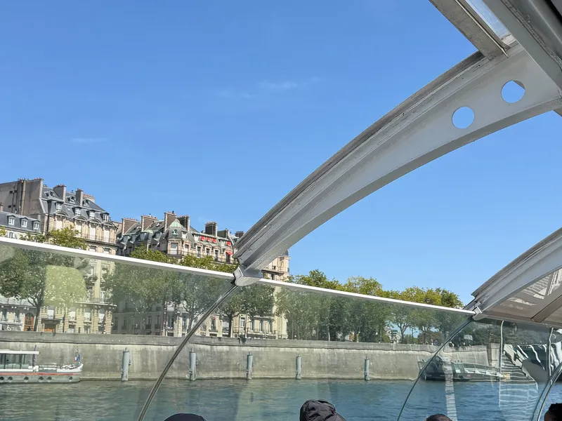 The view from under a curved glass boat roof, looking across the Seine at a quay and apartment buildings